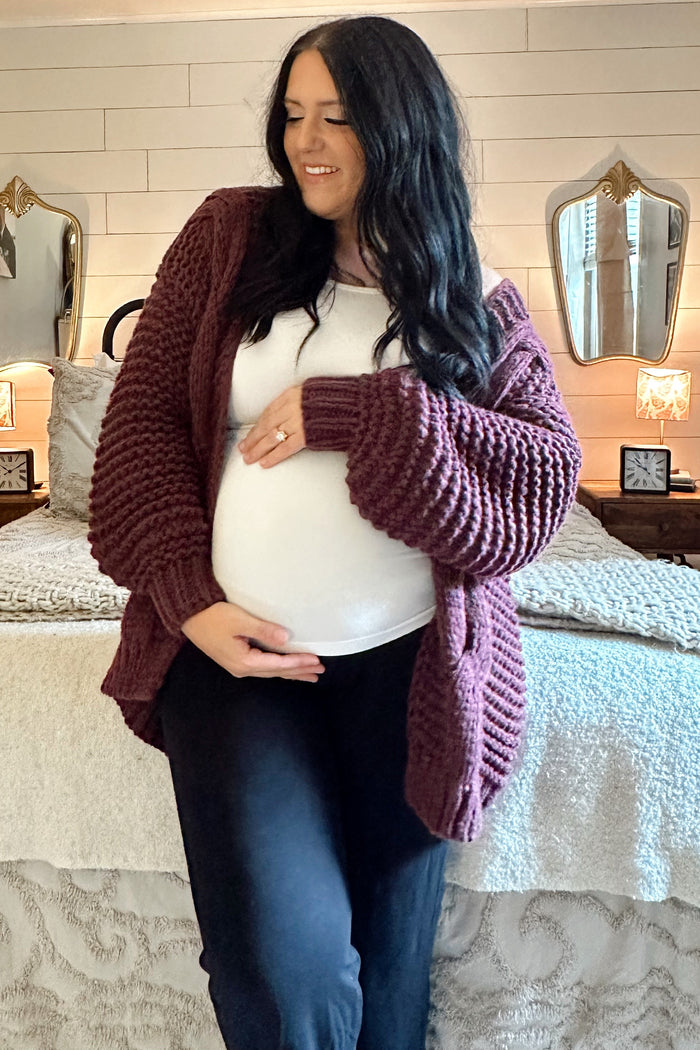 woman wearing a maroon chunky knit cardigan over a white top and holding her baby bump, black pants, in a cozy bedroom setting