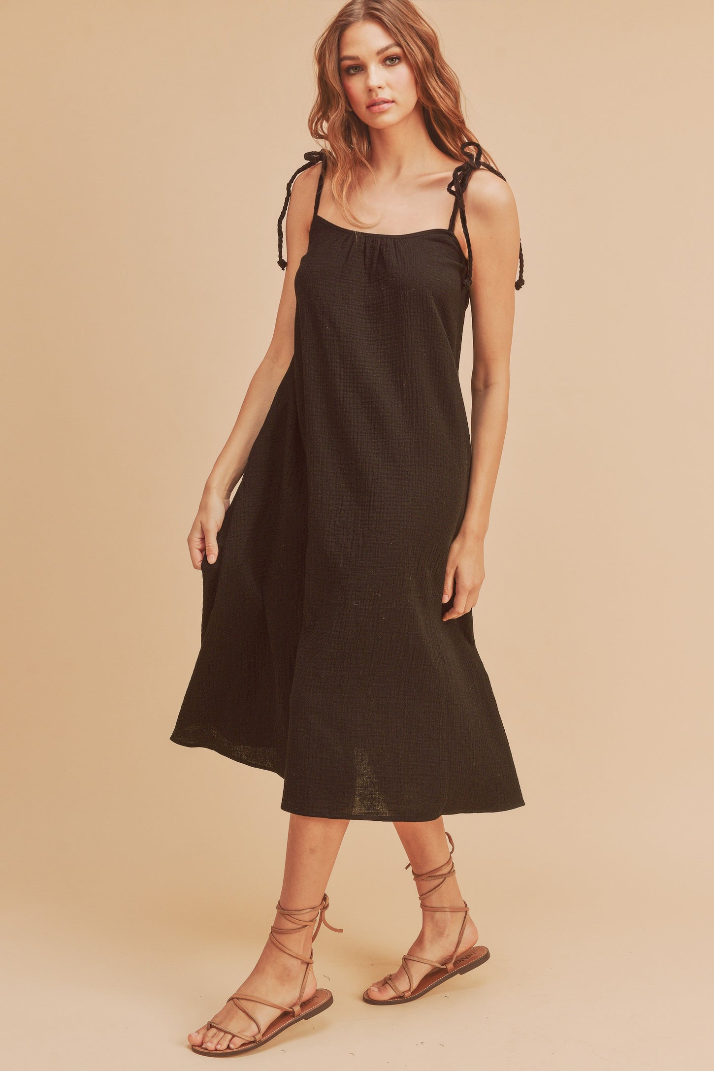 woman wearing a black spaghetti strap midi dress with tie detail on the shoulders and strappy sandals