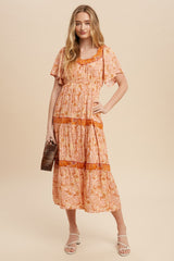 woman wearing a pink and orange floral print maxi dress with short sleeves and tiered ruffle details, holding a brown clutch