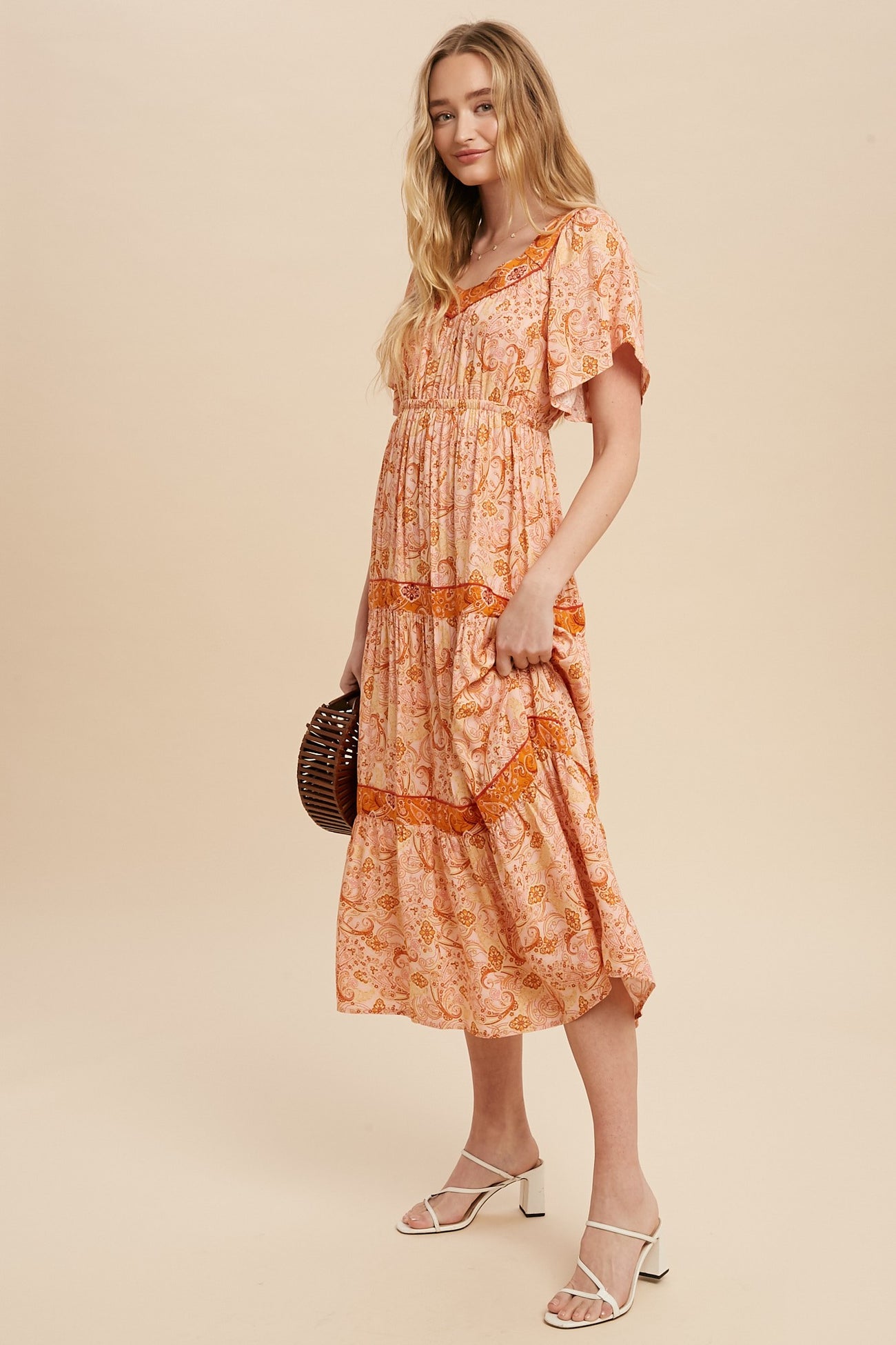 woman wearing a light orange floral maxi dress with short sleeves and tiered detailing, standing with one hand in pocket