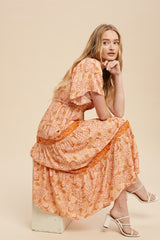 woman wearing a peach floral maxi dress with short sleeves and tiered skirt, sitting on a pedestal
