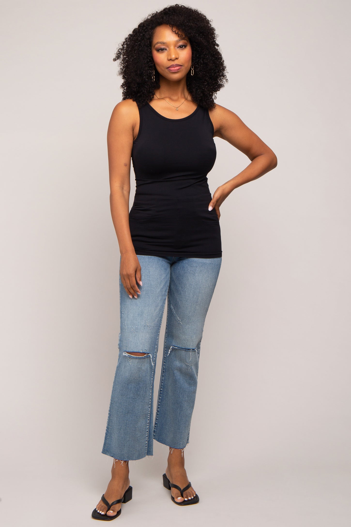 woman wearing a black sleeveless top and distressed light wash flared jeans, standing confidently with one hand on her hip