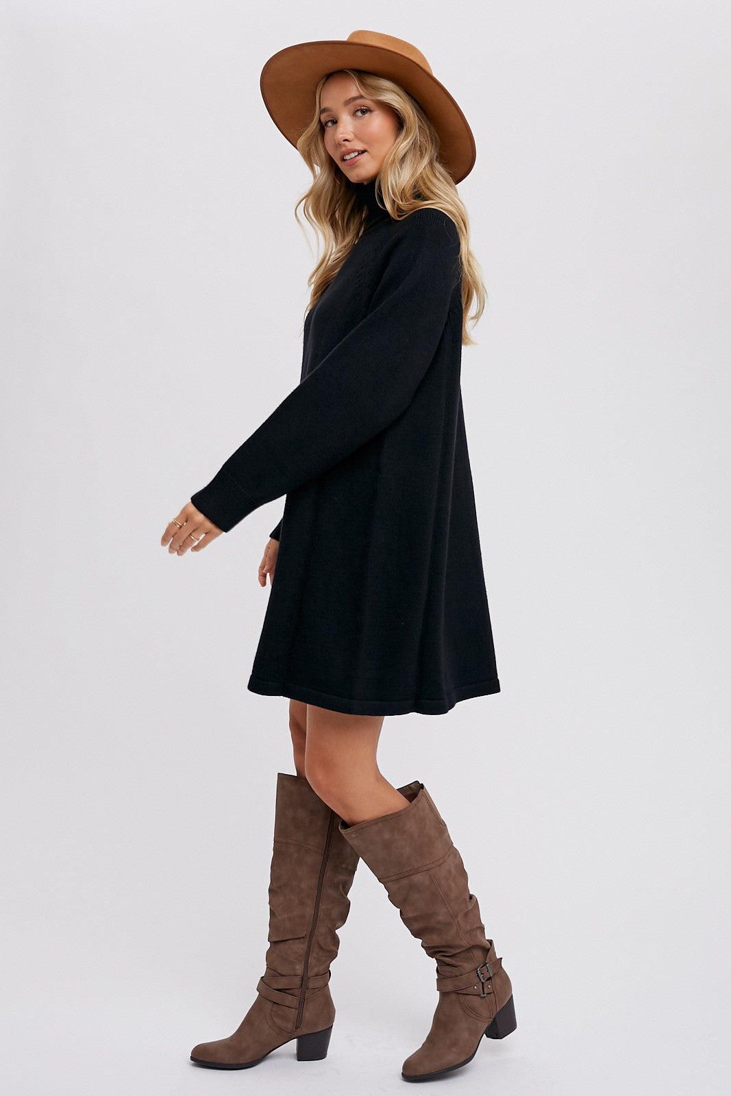 woman wearing a black long sleeve sweater dress, tall brown boots, and a wide-brimmed hat, standing sideways