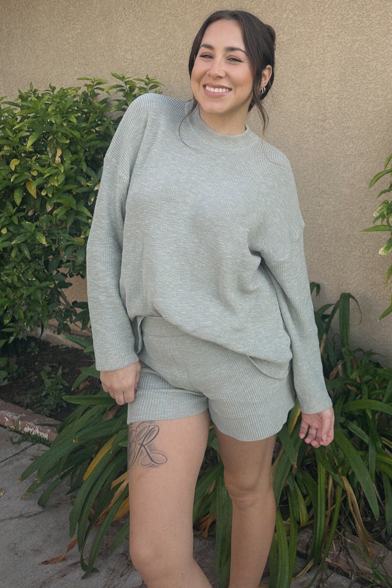 woman wearing a light gray ribbed sweater and matching shorts, smiling outdoors among greenery