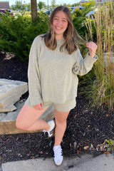 woman wearing a light gray oversized sweater and matching shorts, posing outdoors with a smile