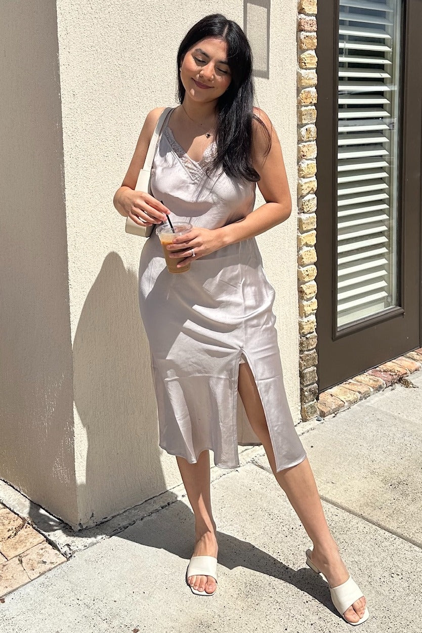 woman wearing a light beige satin slip dress with a side slit, holding a drink and wearing white sandals