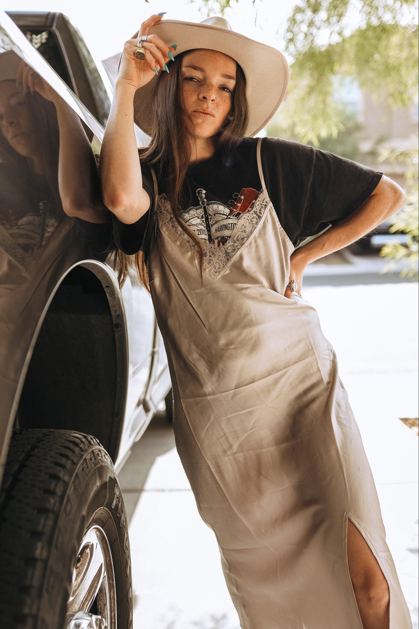woman wearing a beige satin slip dress with lace details and a black graphic t-shirt, posing next to a vehicle
