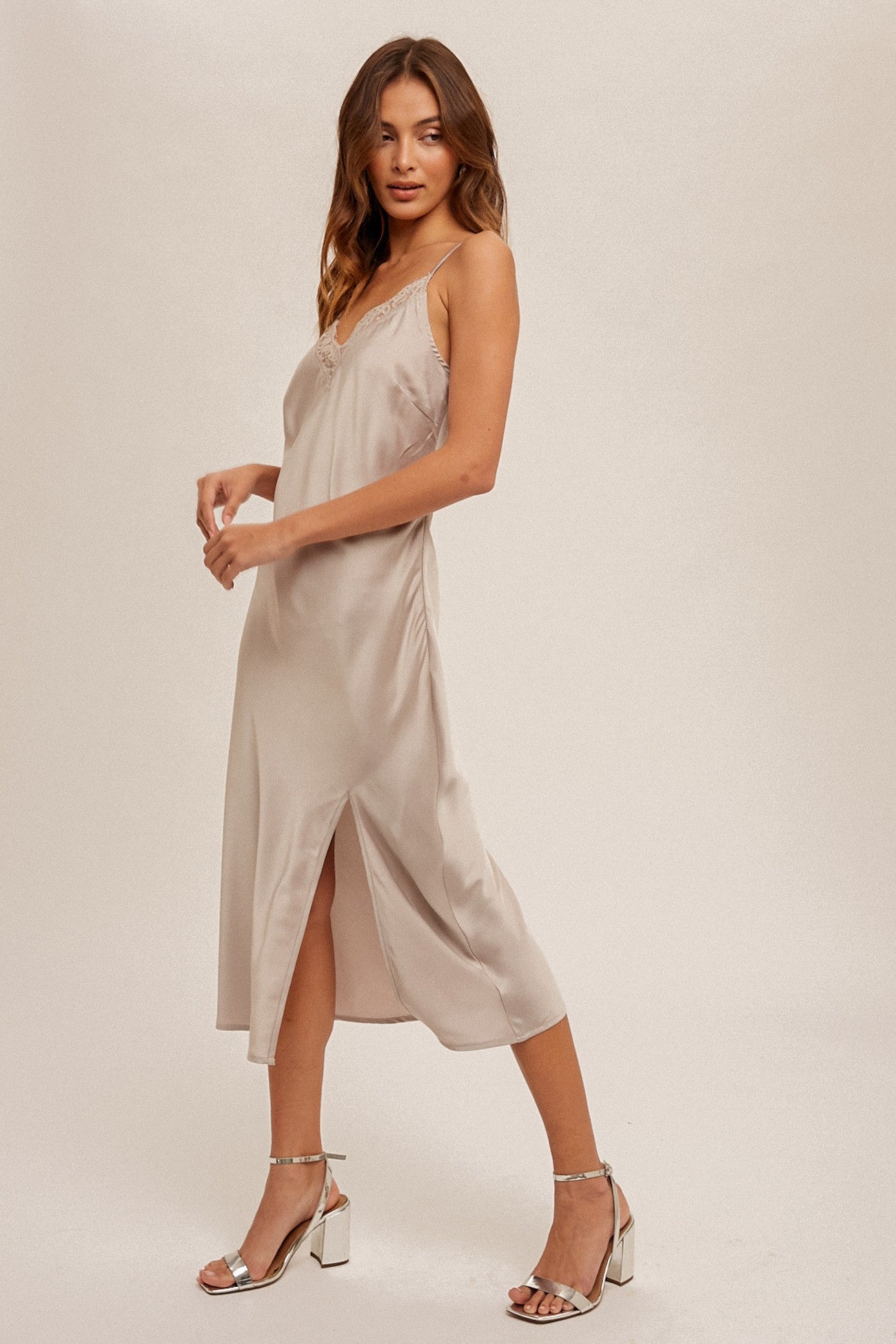 woman wearing a beige satin midi dress with a side slit and thin spaghetti straps, styled with silver high heels