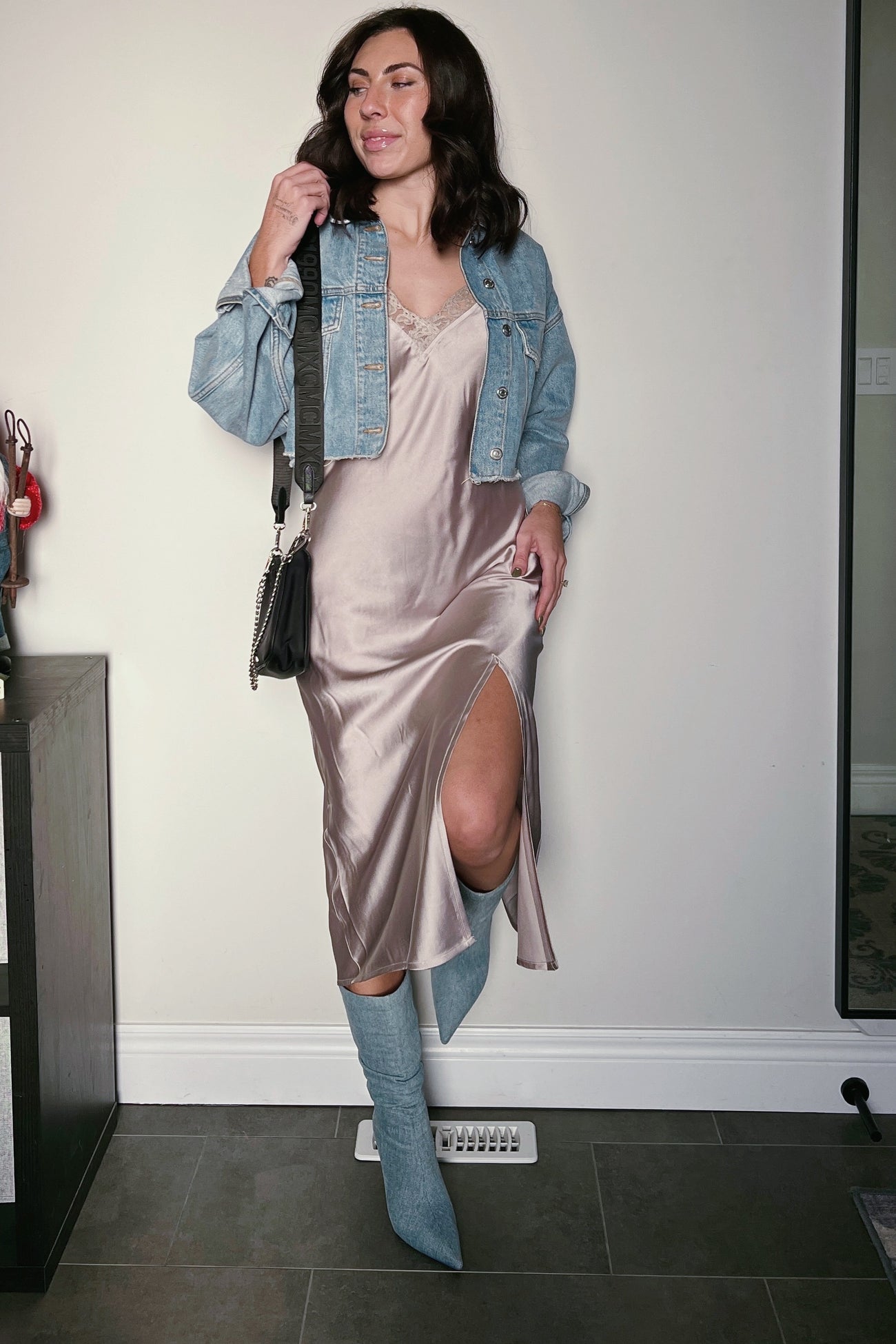 woman wearing a silky taupe slip dress with a thigh slit, denim jacket, and knee-high blue boots, holding a black handbag
