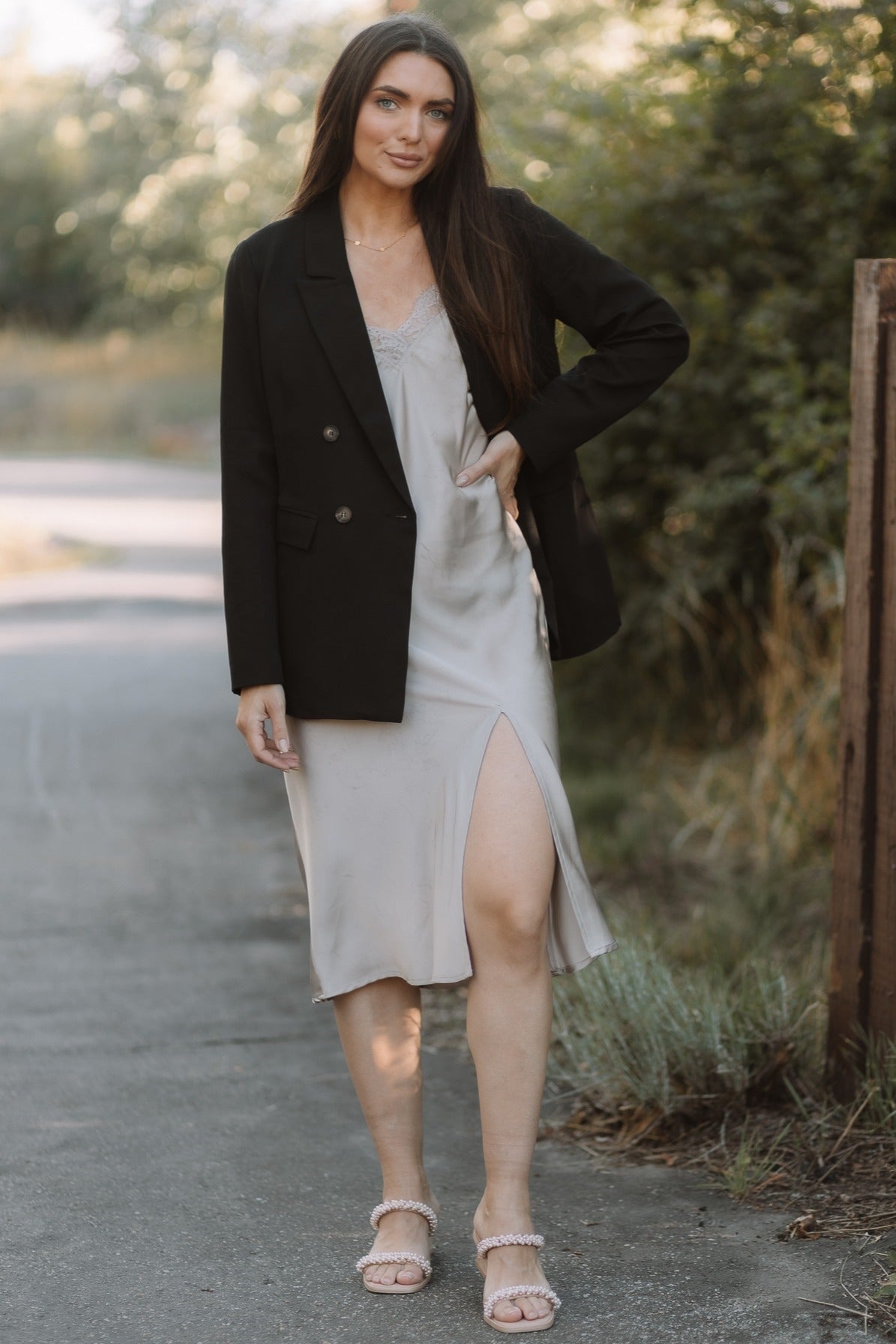 woman wearing a light gray slip dress with a high slit and a black double-breasted blazer, standing outdoors