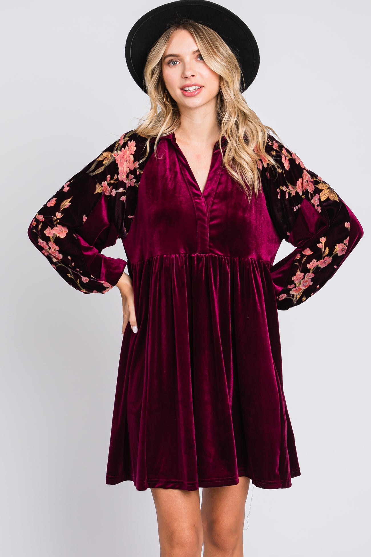 woman wearing a burgundy velvet dress with floral embroidered long sleeves and a v-neckline, posing with hands on hips