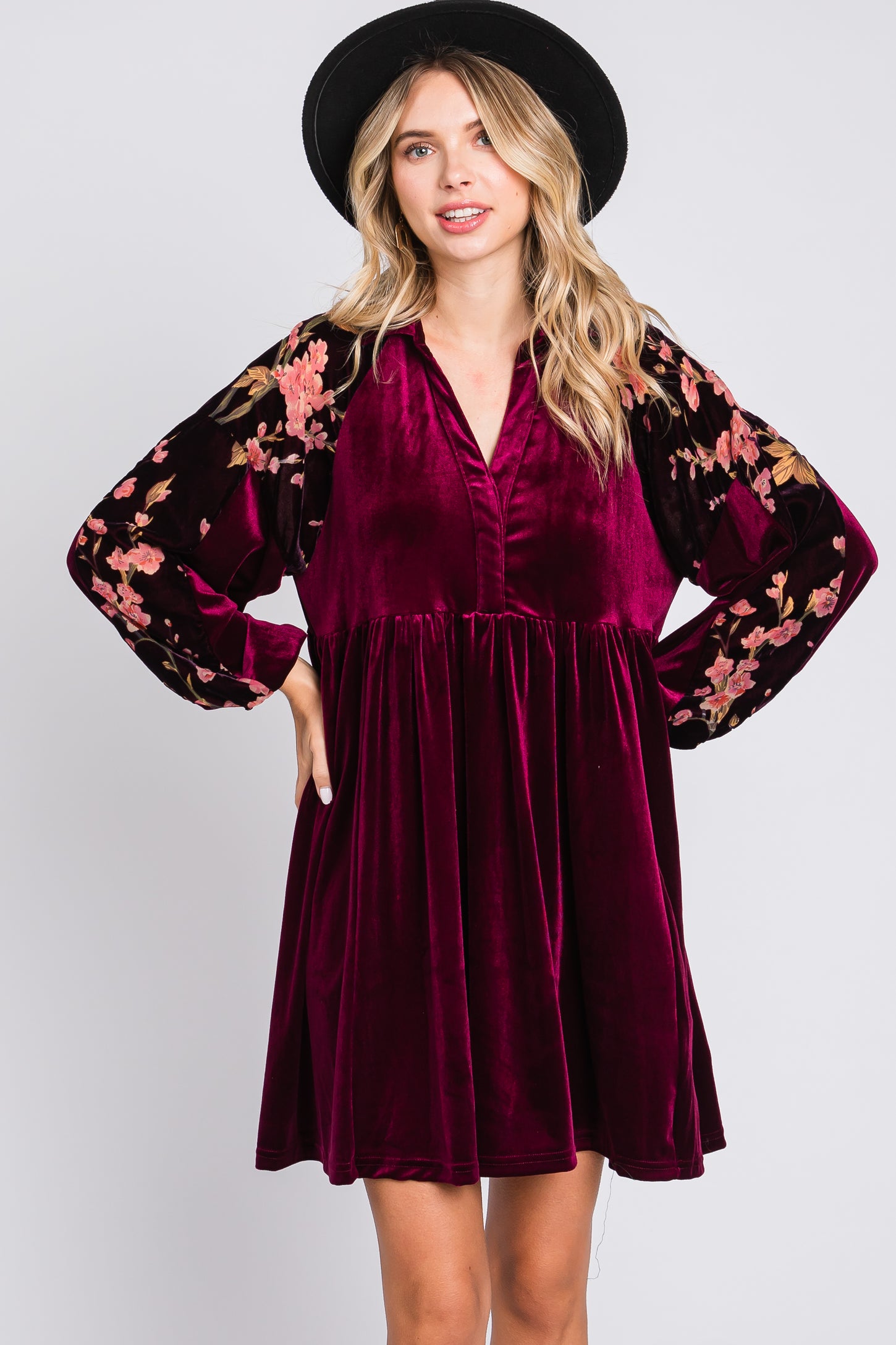 woman wearing a burgundy velvet dress with floral embroidered long sleeves and a v-neckline, posing with hands on hips
