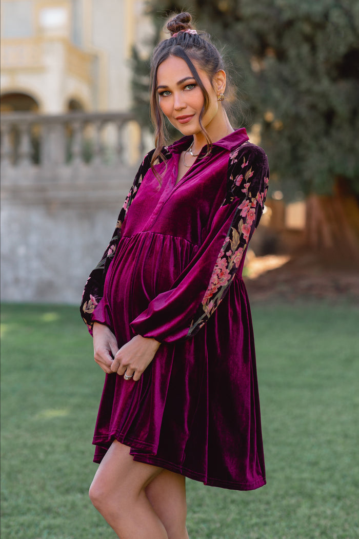 woman wearing a purple velvet maternity dress with floral long sleeves and cradling her baby bump