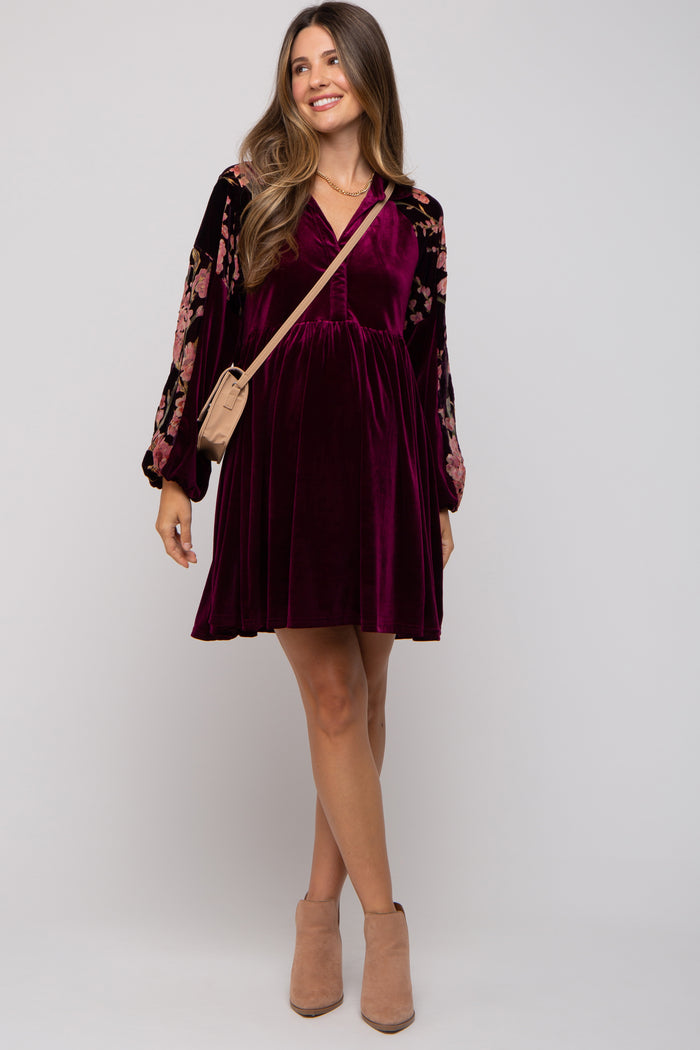 woman wearing a burgundy velvet maternity dress with floral embroidered sleeves and a baby bump, carrying a small bag