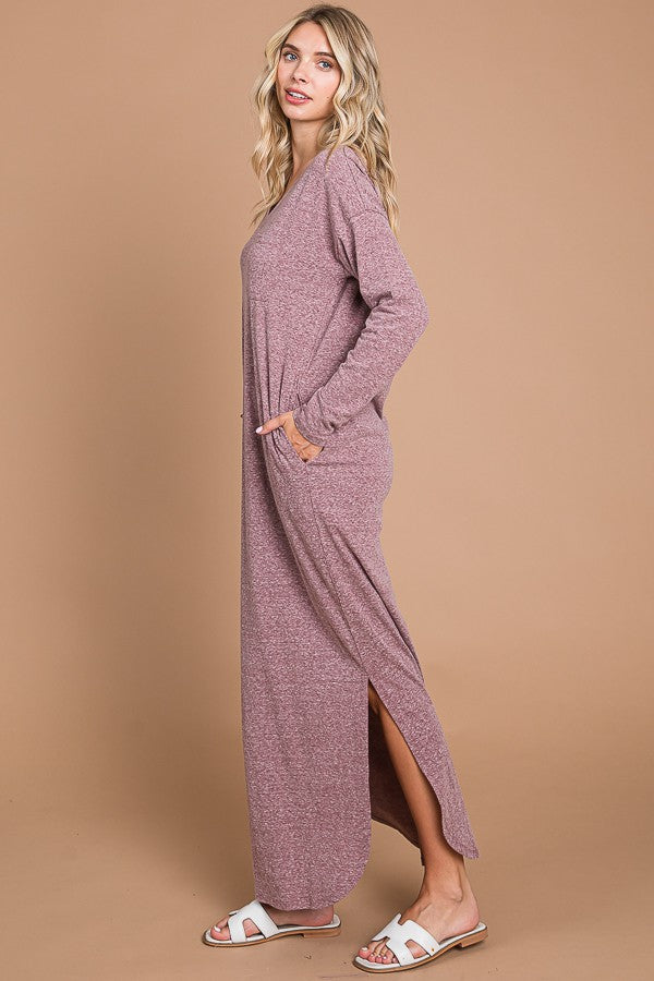 woman wearing a long sleeve mauve maxi dress with side slits and pockets, standing against a light brown background