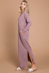woman wearing a long sleeve mauve maxi dress with side slits and pockets, standing against a light brown background