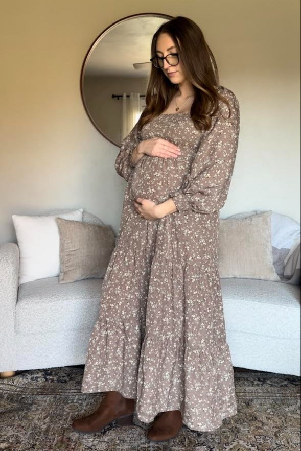 woman wearing a brown floral maternity dress and holding her baby bump while standing in a living room