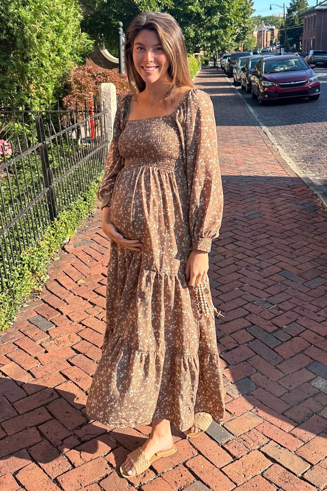 woman wearing a brown floral maternity dress with long sleeves and a tiered skirt, holding her baby bump