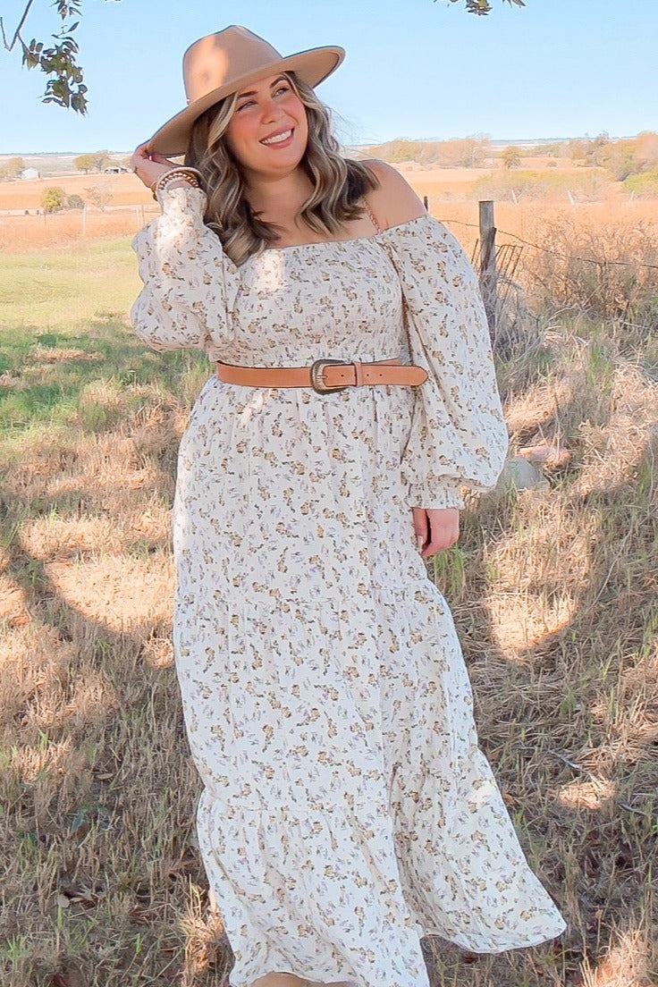 woman wearing a white off-the-shoulder floral maxi dress with long sleeves and a brown belt, accessorized with a wide-brim hat