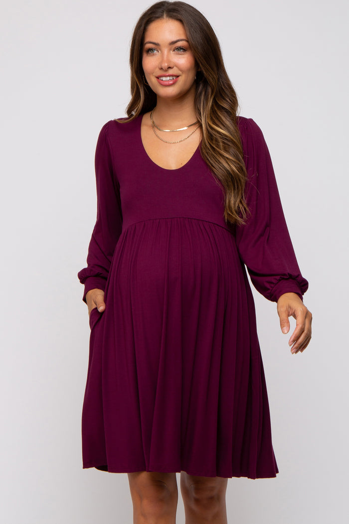 woman wearing a burgundy maternity dress with a v-neck and long sleeves, featuring side pockets and a flowy skirt