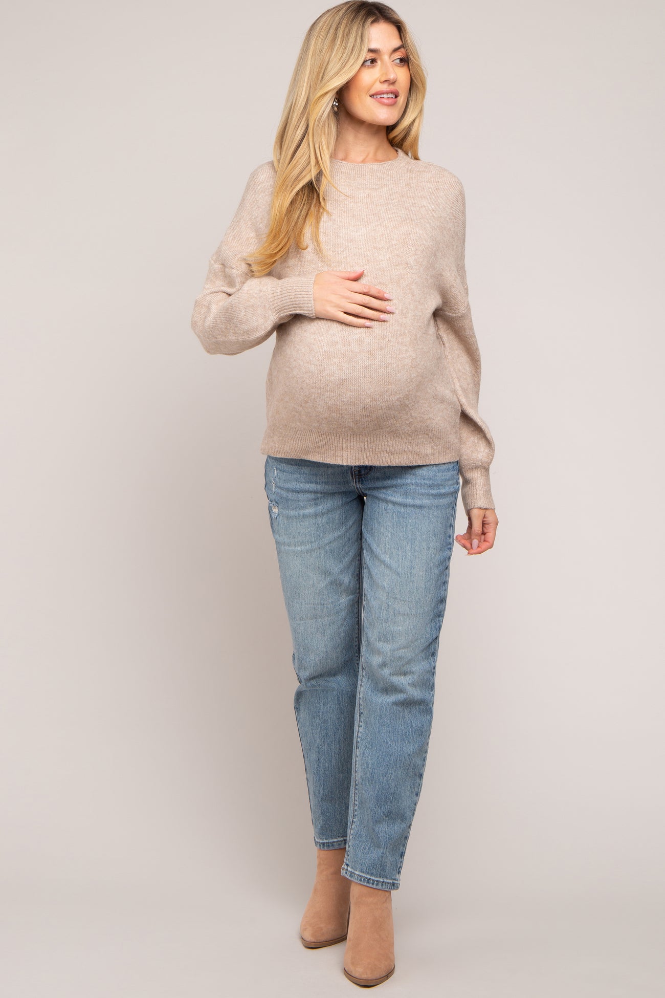 woman wearing a beige maternity sweater and light blue jeans, showing her baby bump with a soft smile