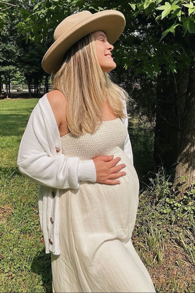 woman wearing a white strapless maternity dress and a cream cardigan, holding her baby bump outdoors