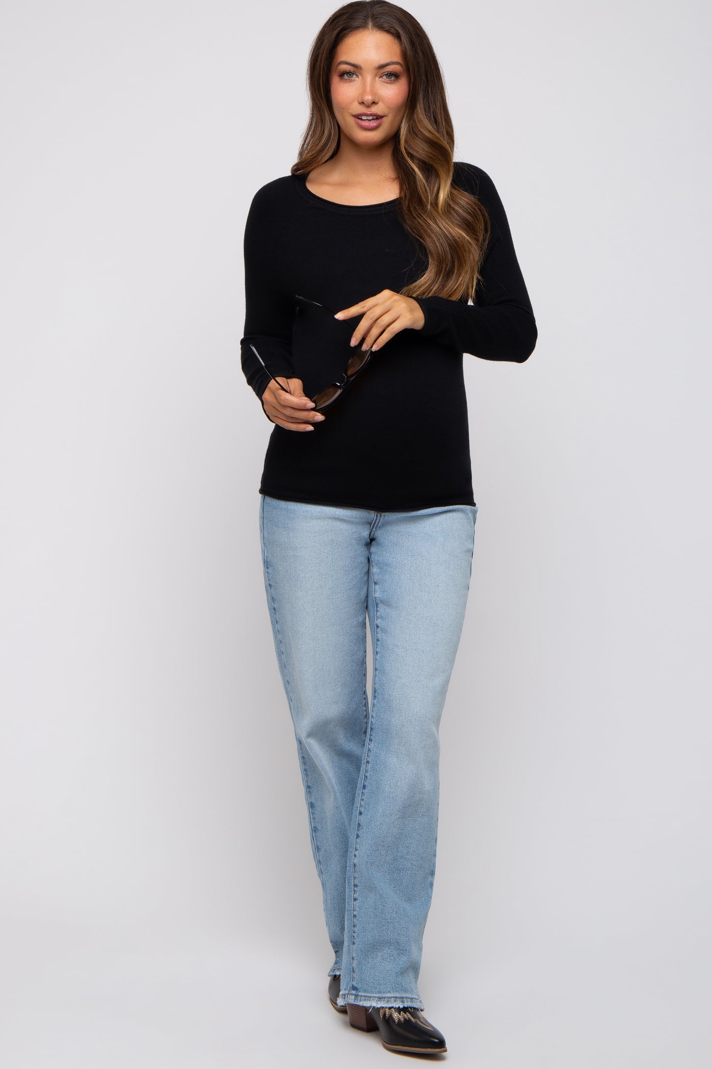 woman wearing a black long sleeve maternity top and light blue flared jeans, holding sunglasses