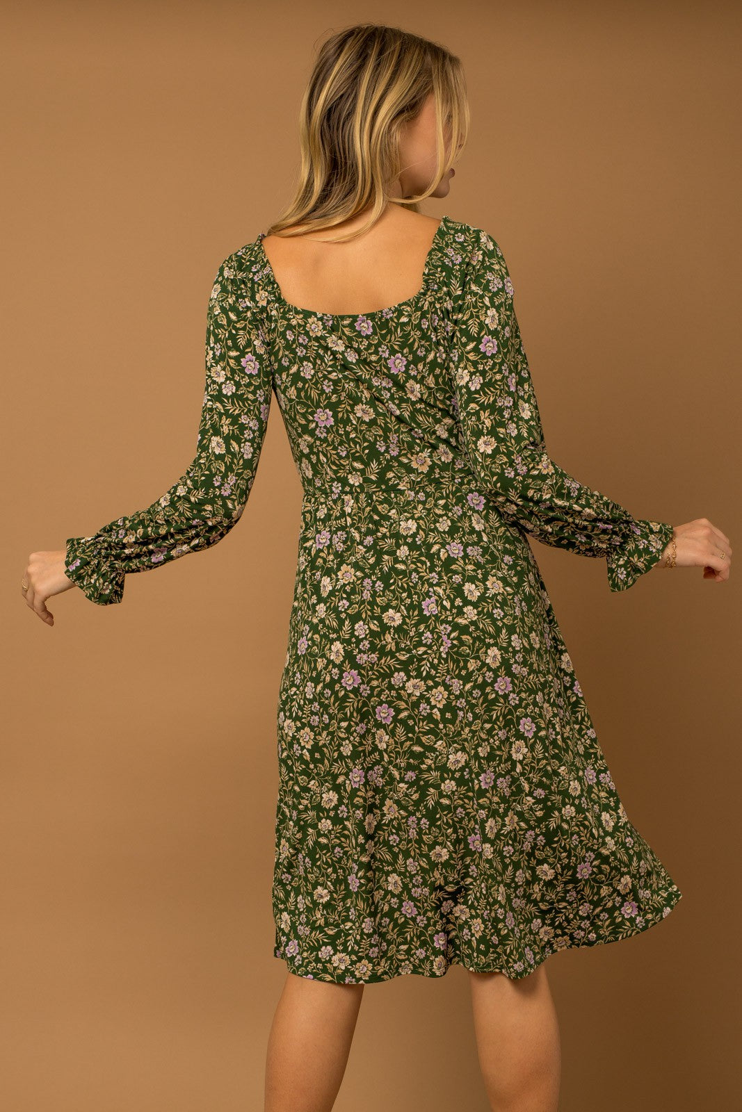 woman wearing a green floral long sleeve dress with a square neckline and flared cuffs, seen from the back