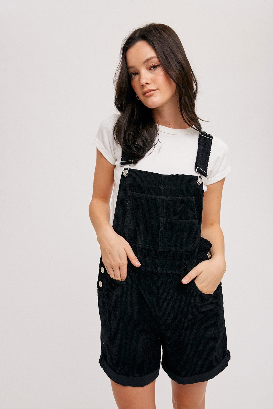 woman wearing a black corduroy overall shorts with a white t-shirt underneath and hands in pockets