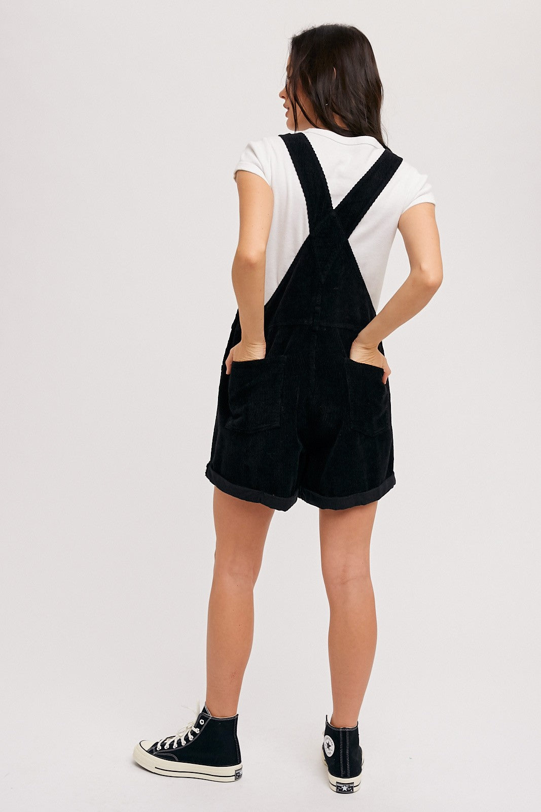 woman wearing black corduroy overall shorts with a white t-shirt, turned back to show the straps and pockets
