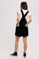 woman wearing black corduroy overall shorts with a white t-shirt, turned back to show the straps and pockets