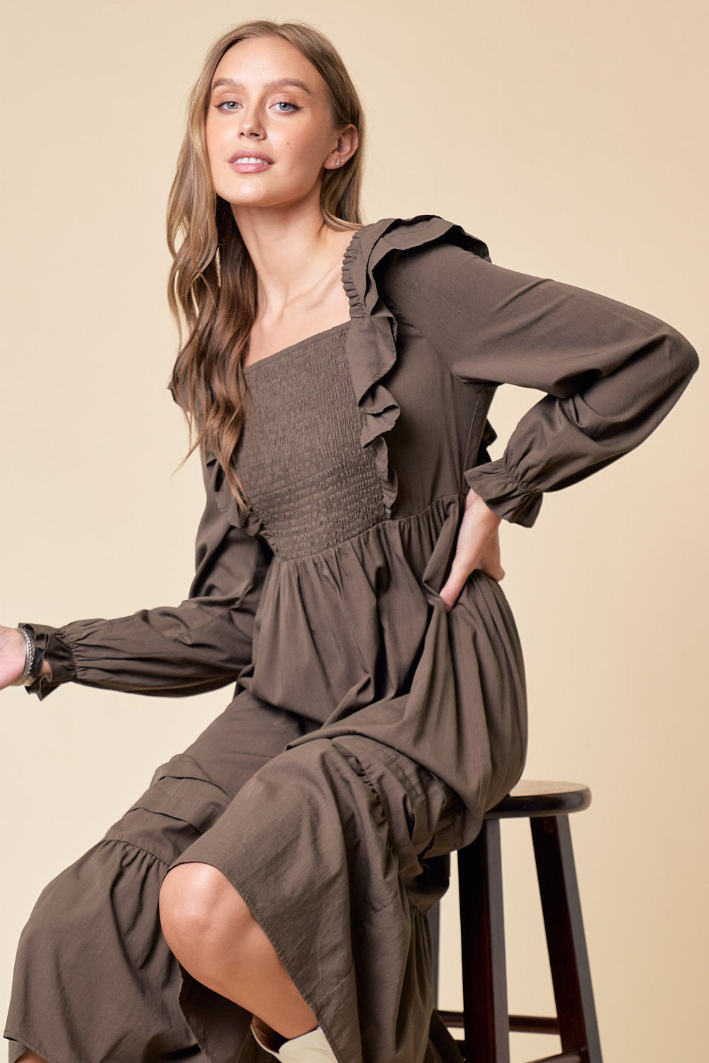 woman wearing an olive green long sleeve dress with ruffle details and a smocked bodice, seated on a stool
