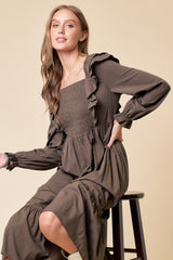 woman wearing an olive green long sleeve dress with ruffle details and a smocked bodice, seated on a stool