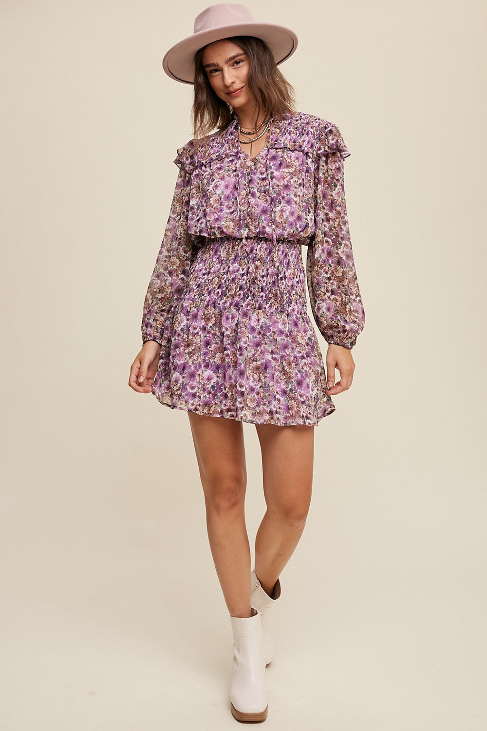 woman wearing a purple floral mini dress with ruffle details and long sleeves, paired with a pink hat and white ankle boots