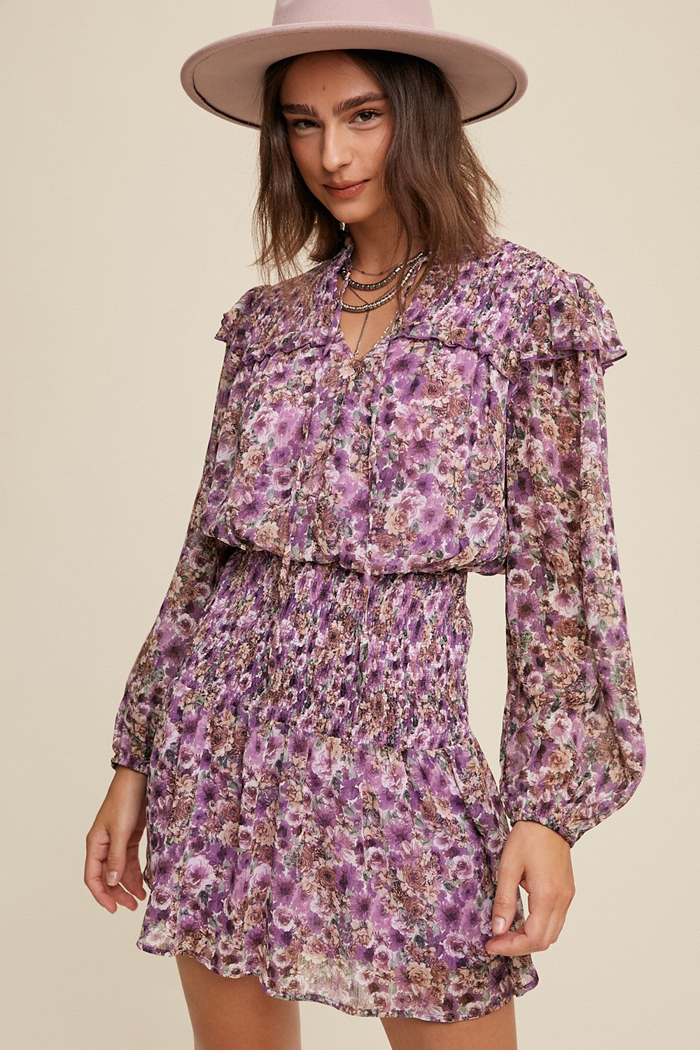 woman wearing a purple floral smocked mini dress with ruffle details and long sleeves, paired with a wide-brimmed hat