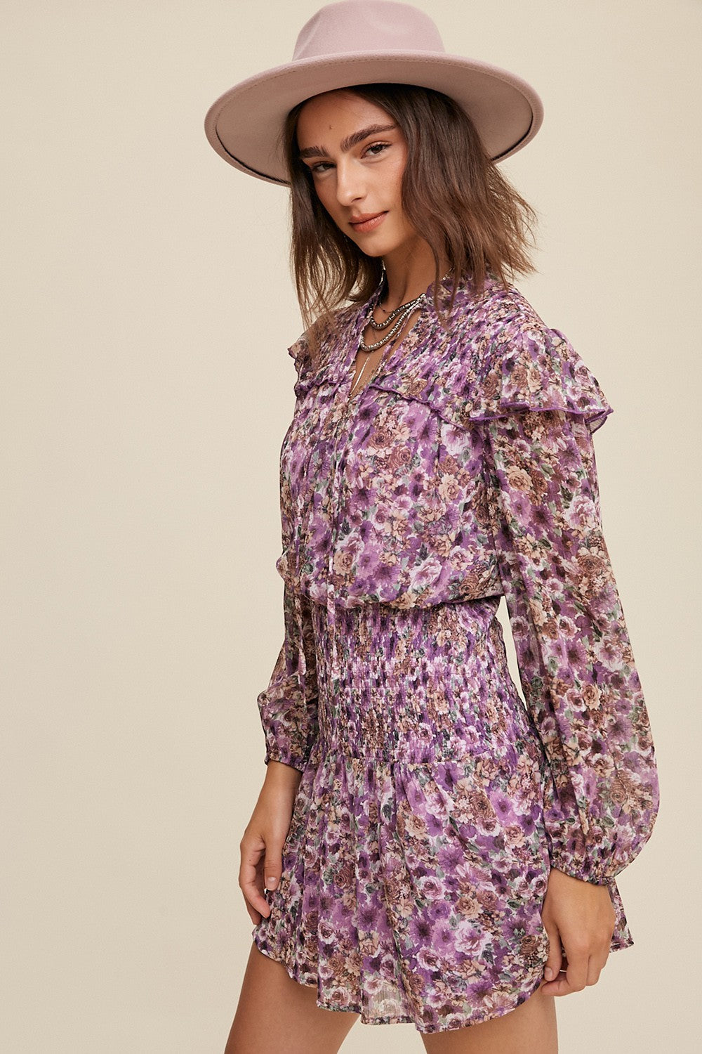 woman wearing a purple floral ruffled mini dress with a smocked waist and long sleeves, accessorized with a wide-brimmed hat
