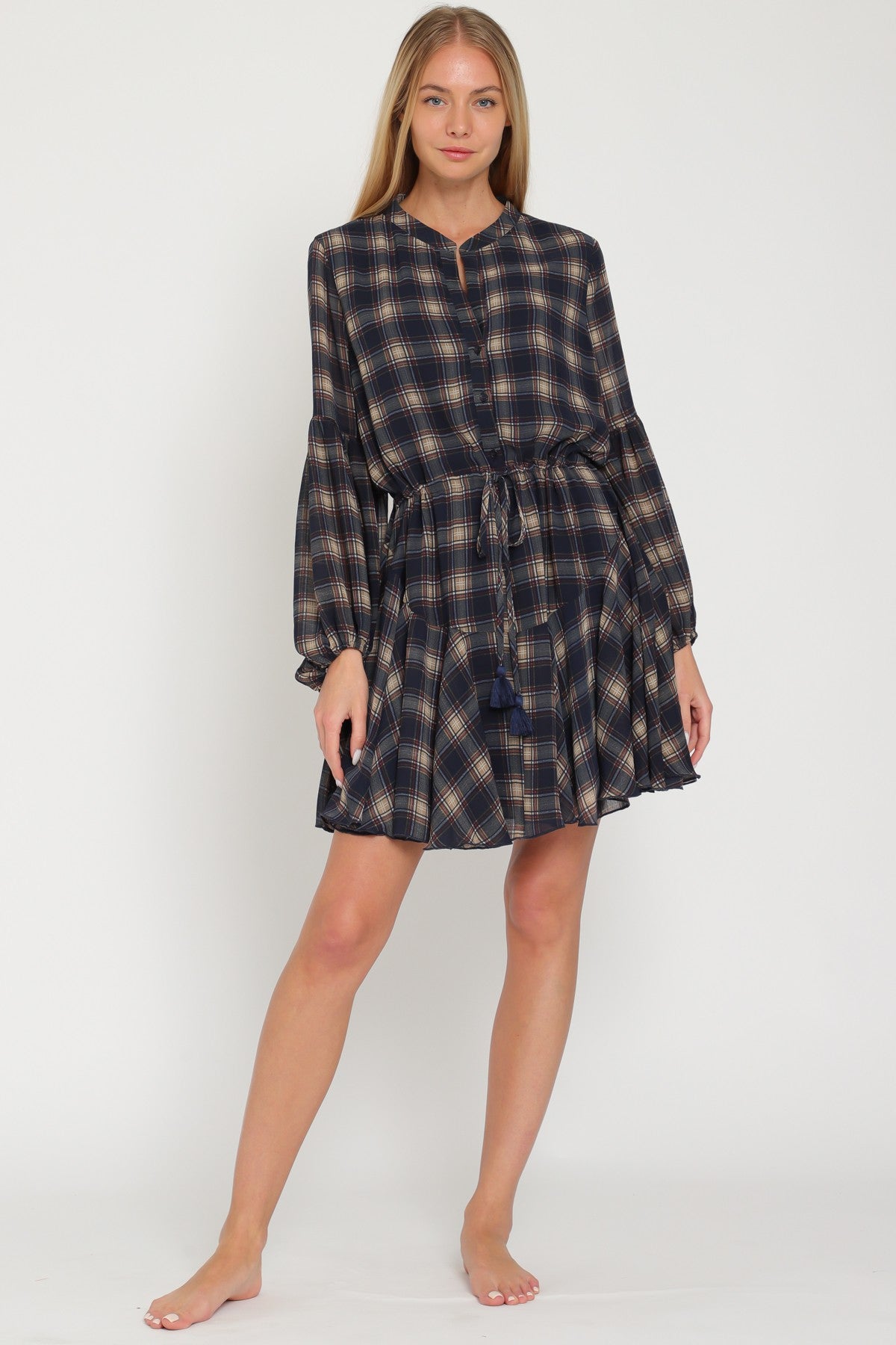 woman wearing a navy and beige plaid short dress with long sleeves and a tie waist detail, standing barefoot