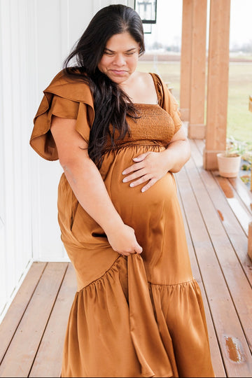 woman wearing a brown smocked maternity dress with ruffle sleeves and holding her baby bump