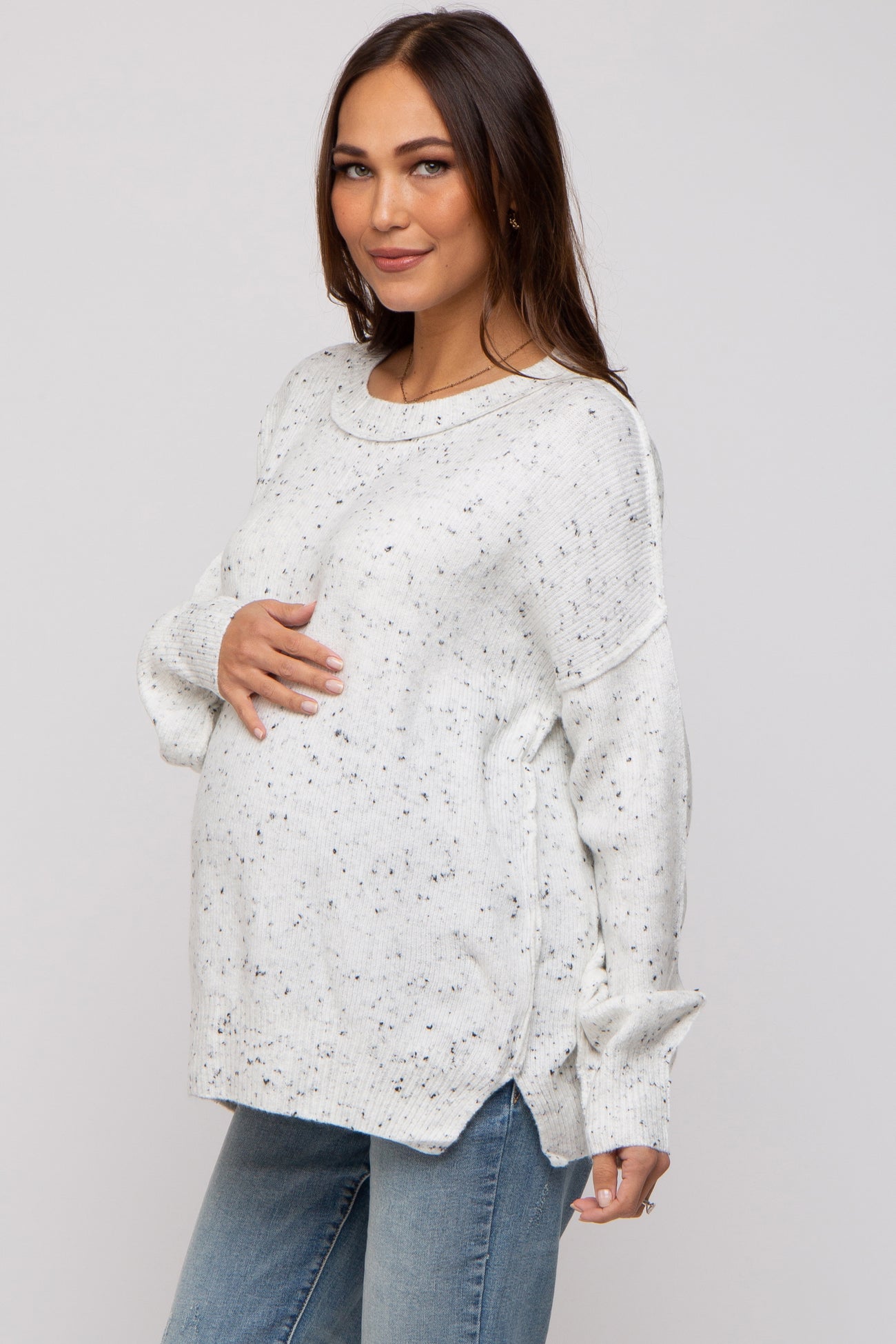 woman wearing a cream speckled maternity sweater with side slits and holding her baby bump