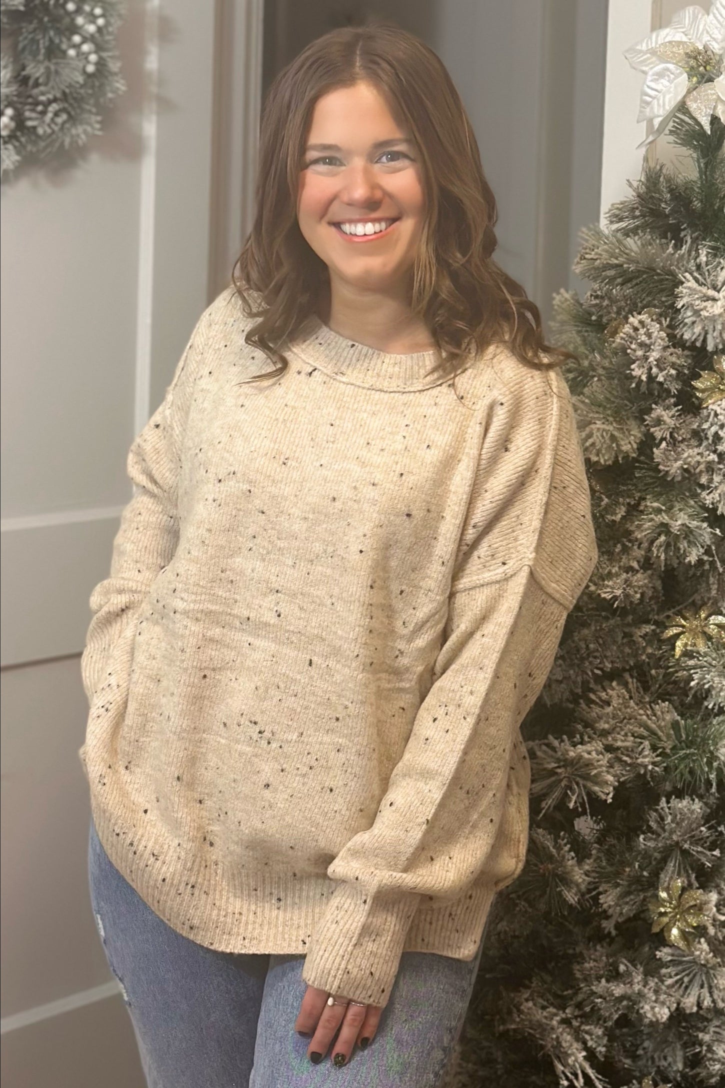 woman wearing a beige speckled oversized sweater and light blue jeans, standing in front of a Christmas tree