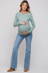 woman wearing a light green ribbed maternity sweater and denim flare jeans, holding her baby bump