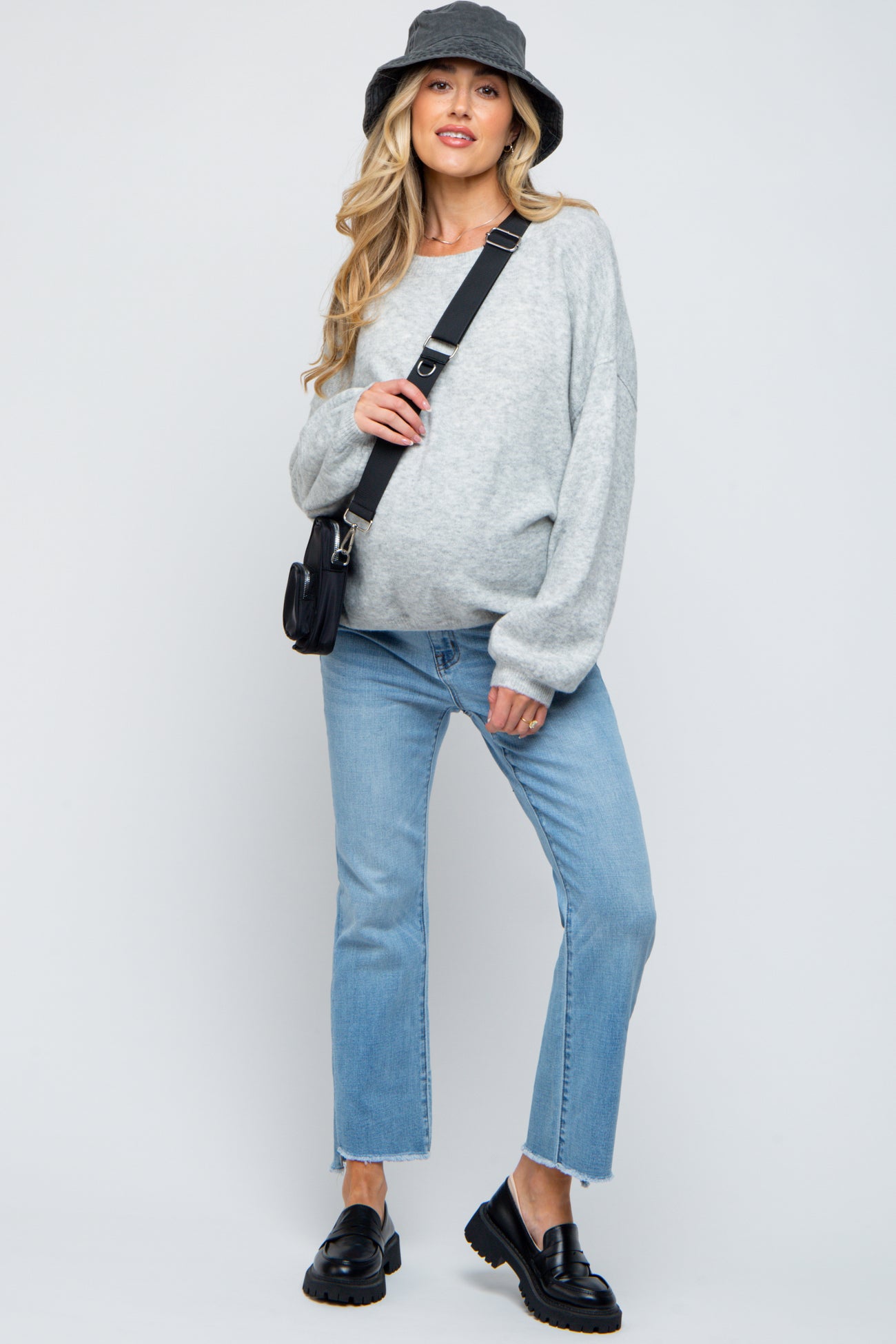 woman wearing a gray oversized sweater and light blue jeans with a baby bump, accessorized with a black bucket hat and bag