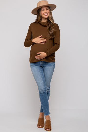 woman wearing a brown long sleeve maternity top and blue skinny jeans, holding her baby bump, wearing a hat and sandals