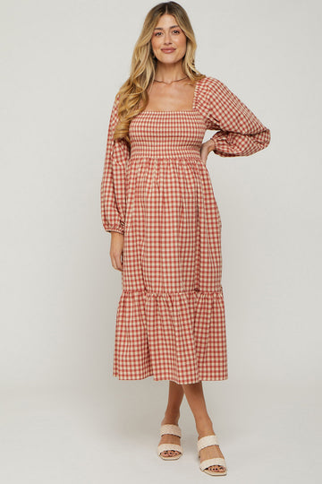 woman wearing a pink and white gingham maternity dress with long sleeves and a tiered hem, holding her baby bump