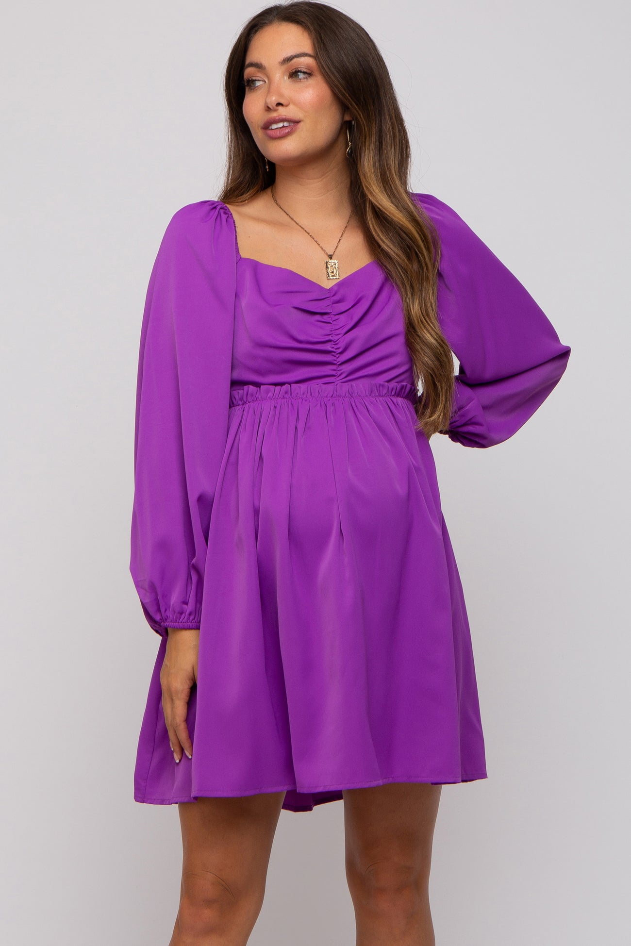 woman wearing a purple maternity dress with long puff sleeves and a ruched bodice accentuating her baby bump