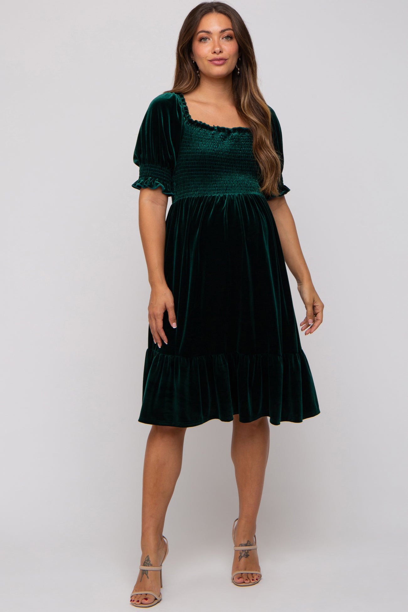 woman wearing a dark green velvet maternity dress with short puff sleeves and a smocked bodice, featuring a ruffled hemline