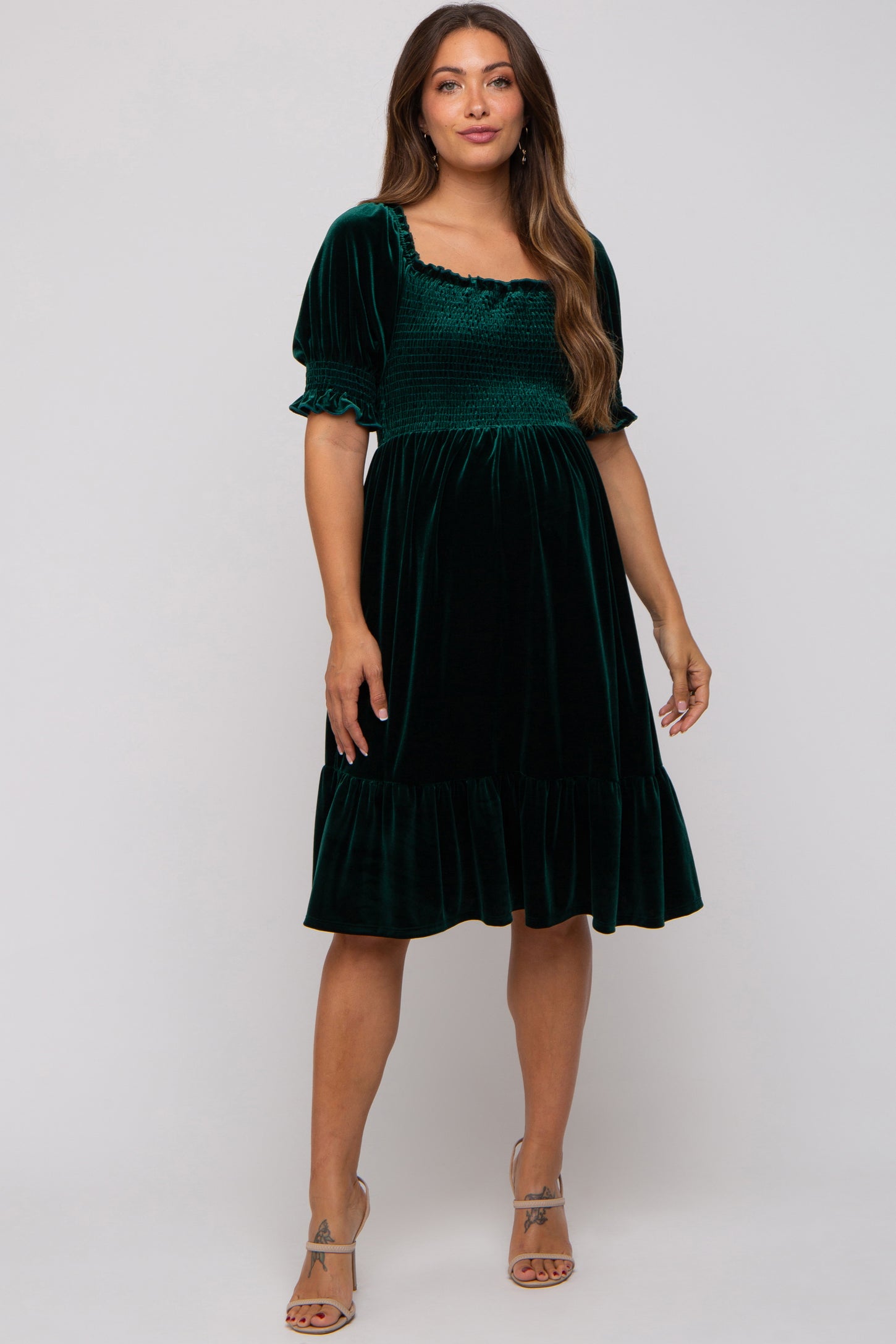 woman wearing a dark green velvet maternity dress with short puff sleeves and a smocked bodice, featuring a ruffled hemline