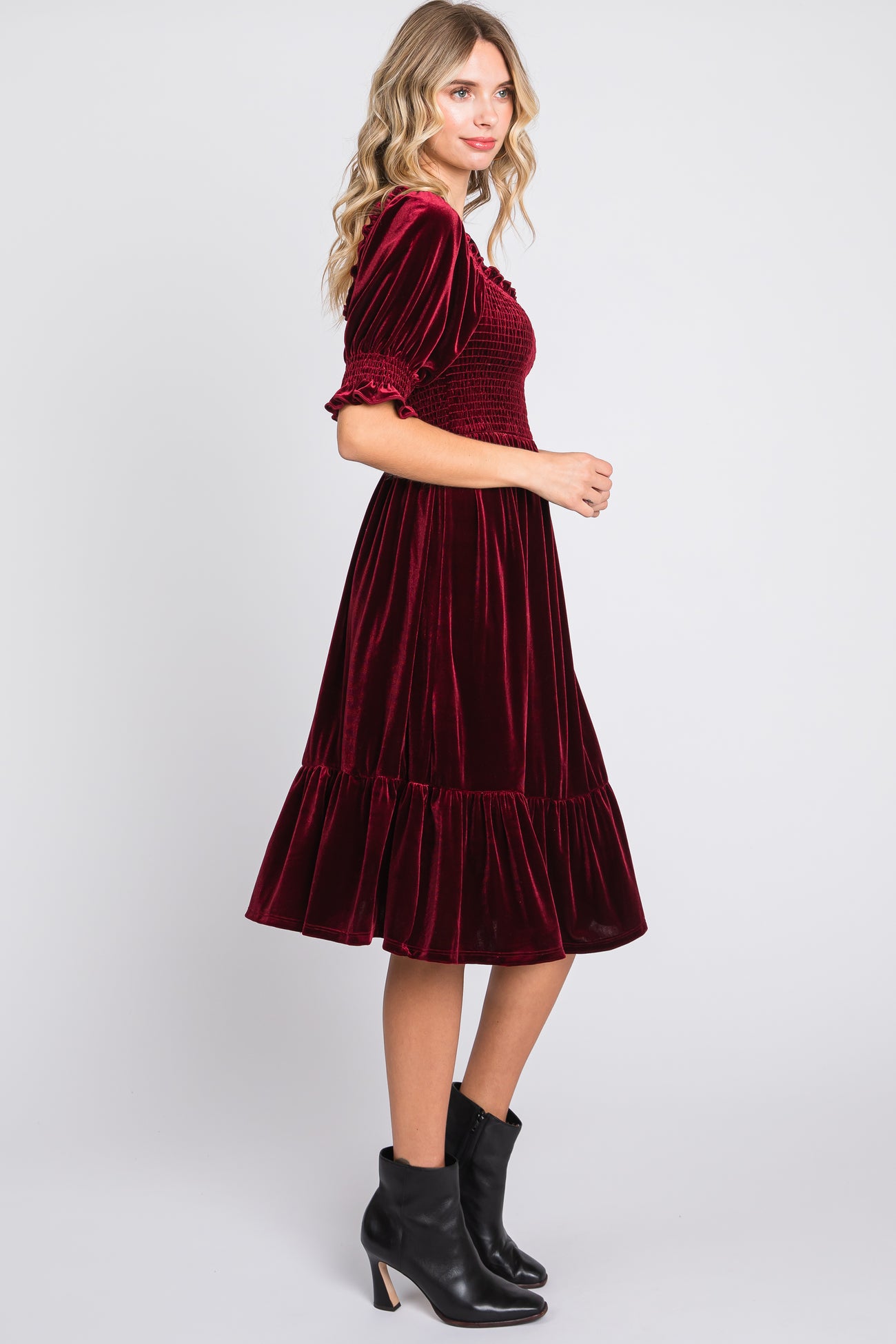 woman wearing a burgundy velvet midi dress with smocked bodice and ruffled hem, paired with black ankle boots