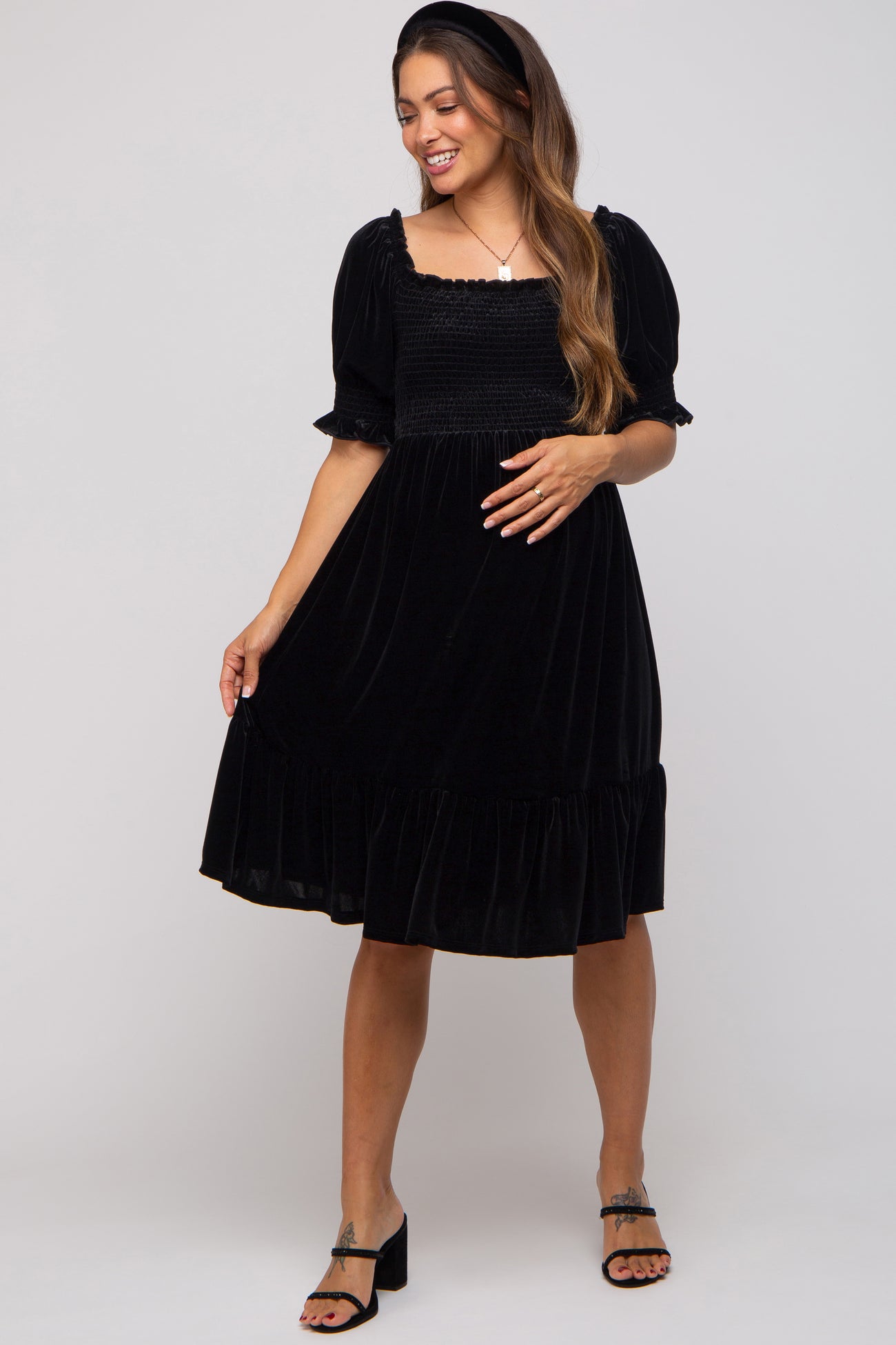 woman wearing a black velvet maternity dress with puff sleeves and a ruffled hem, posing with one hand on her hip