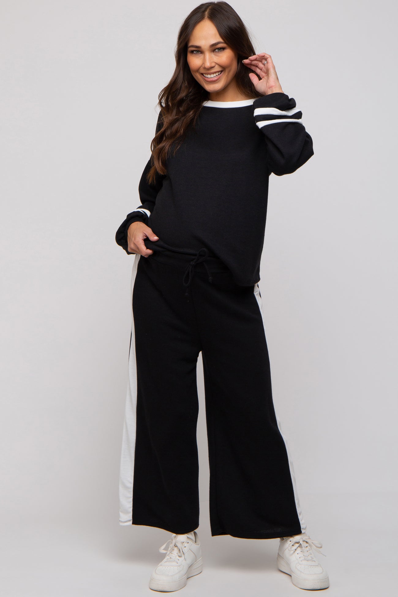 woman wearing a black maternity sweatshirt with white stripes and black wide-leg pants, smiling and posing with hands on her hips